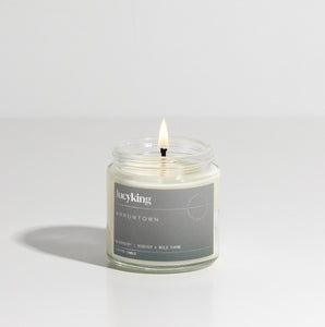 Small best selling Arrowtown soy wax scented candle with a burning wick in a clear glass jar with grey label 